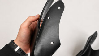 Hand holding a black curved mounting bracket with three holes for screws against a light background